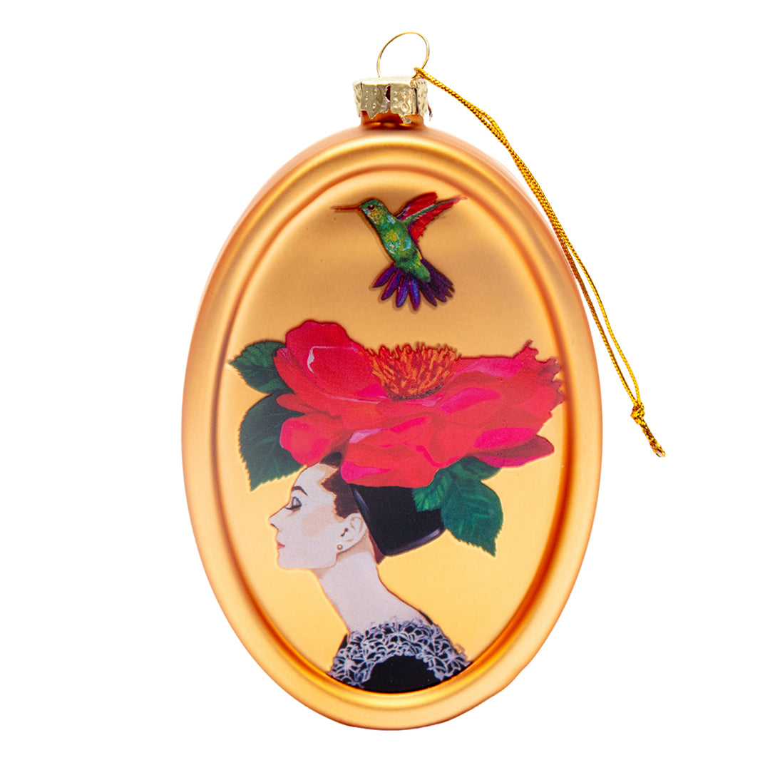 Front view of Ashley Longshore’s hand-blown glass ornament featuring a woman in profile with a large red flower hat and a colorful hummingbird above her head on a gold oval background.