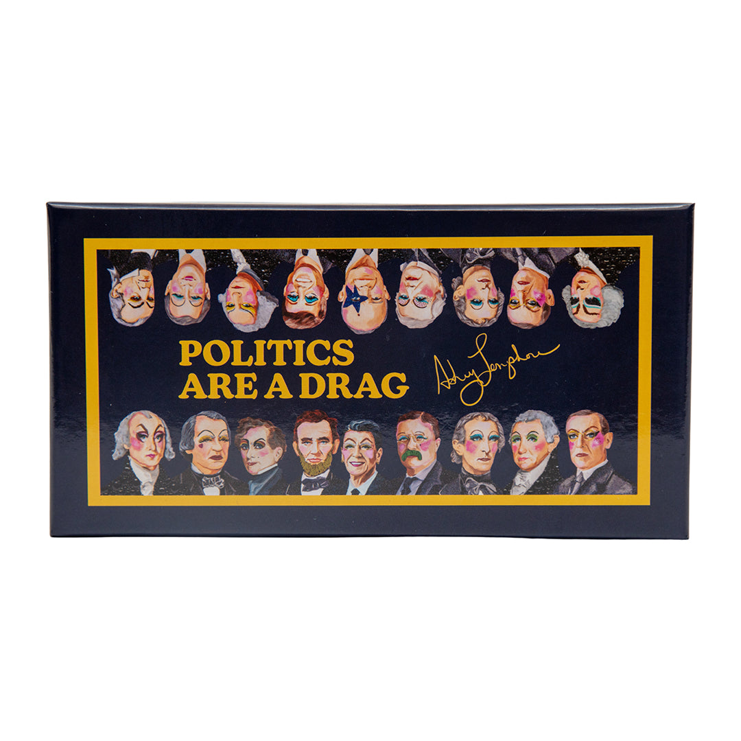 A navy and gold limited-edition box of Ashley Longshore’s Politics Are a Drag Brain Snatch memory game surrounded by illustrated cards of U.S. presidents painted in colorful drag makeup.
