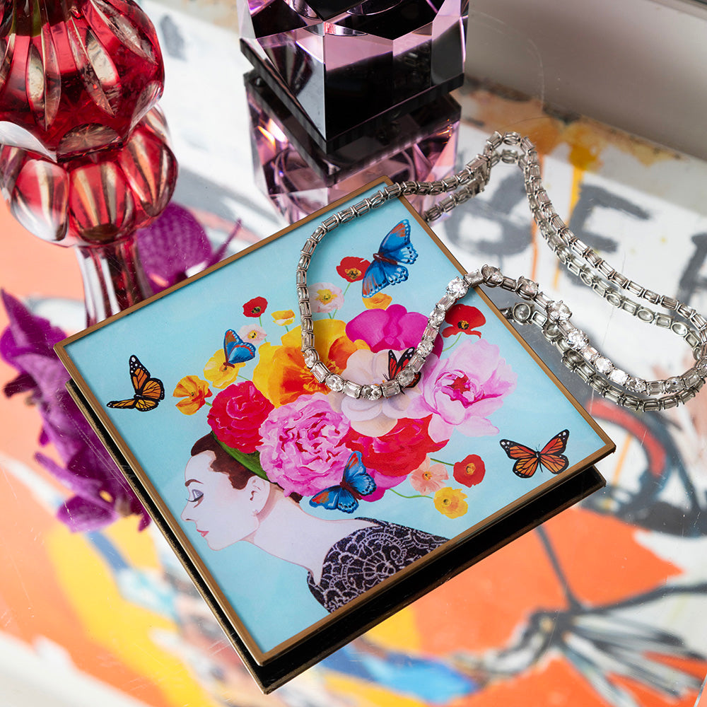 6x6 inch glass deco tray featuring Ashley Longshore’s “Audrey with Flowers and Butterflies” artwork, used as a jewelry catchall. The tray displays a profile of Audrey with a vibrant floral and butterfly crown, placed on a mirrored surface with a crystal necklace and glass décor around it.