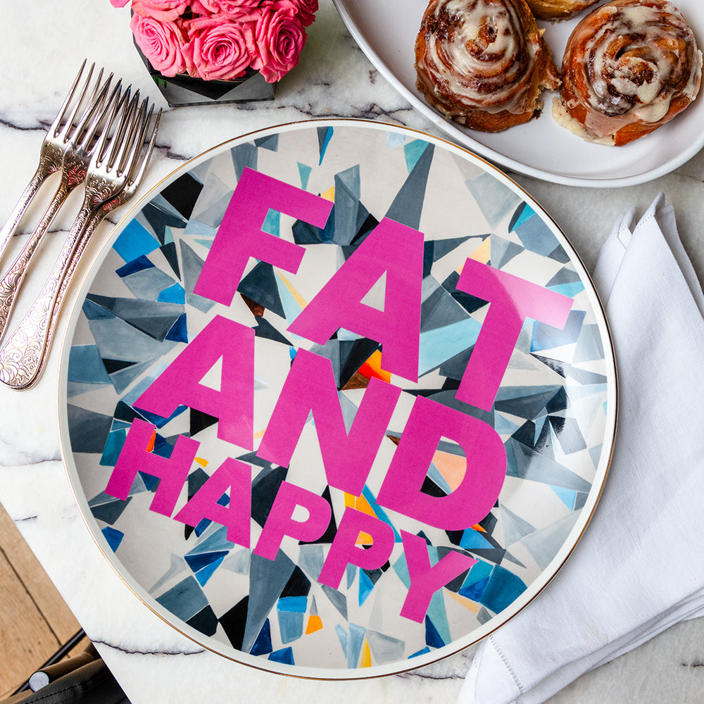 14-inch porcelain platter with the words “FAT AND HAPPY” in bold pink lettering over a geometric diamond-inspired background, styled on a marble table beside silver forks, a bowl of cinnamon rolls, and a small bouquet of pink roses.