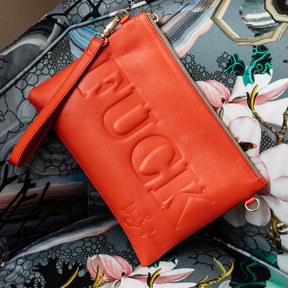 The orange leather bag styled on patterned fabric, showcasing the embossed lettering and wristlet strap.