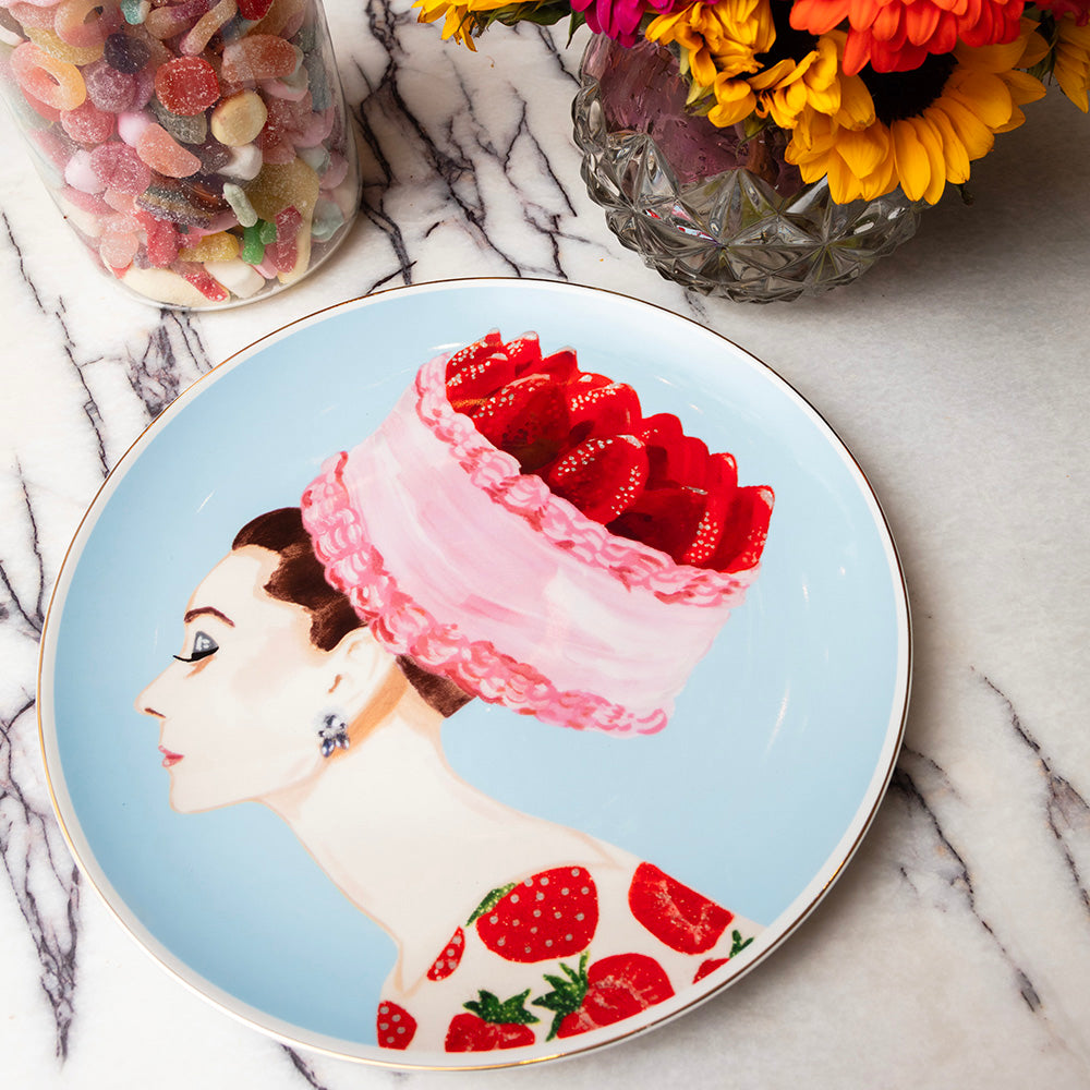 Porcelain “Strawberry Audrey” platter on a marble tabletop, featuring a profile of a woman with a strawberry-topped pink cake hat and strawberry-print dress. Beside the plate is a colorful bouquet of flowers and a glass jar filled with assorted gummy candies.