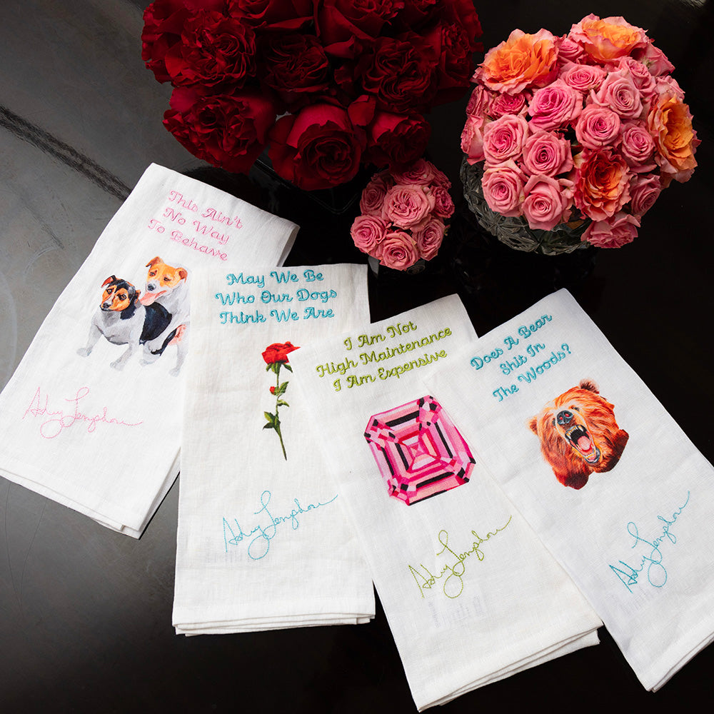 Set of decorative towels with humorous sayings and images, placed on a surface with flowers in the background.
