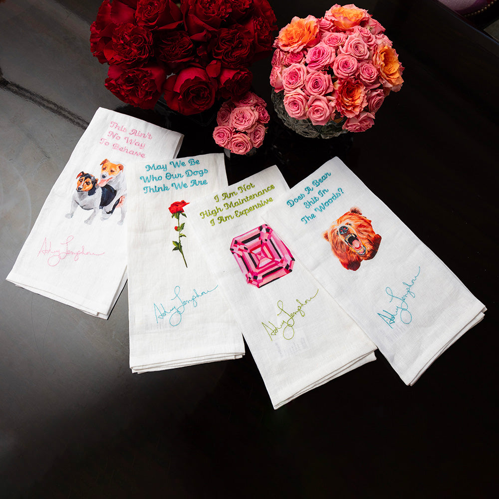 Set of four white linen tea towels laid out on a table, each featuring humorous phrases and artwork by Ashley Longshore — including dogs, a rose, a gemstone, and a bear — with embroidered artist signatures. Pink and red floral arrangements are placed above the towels.