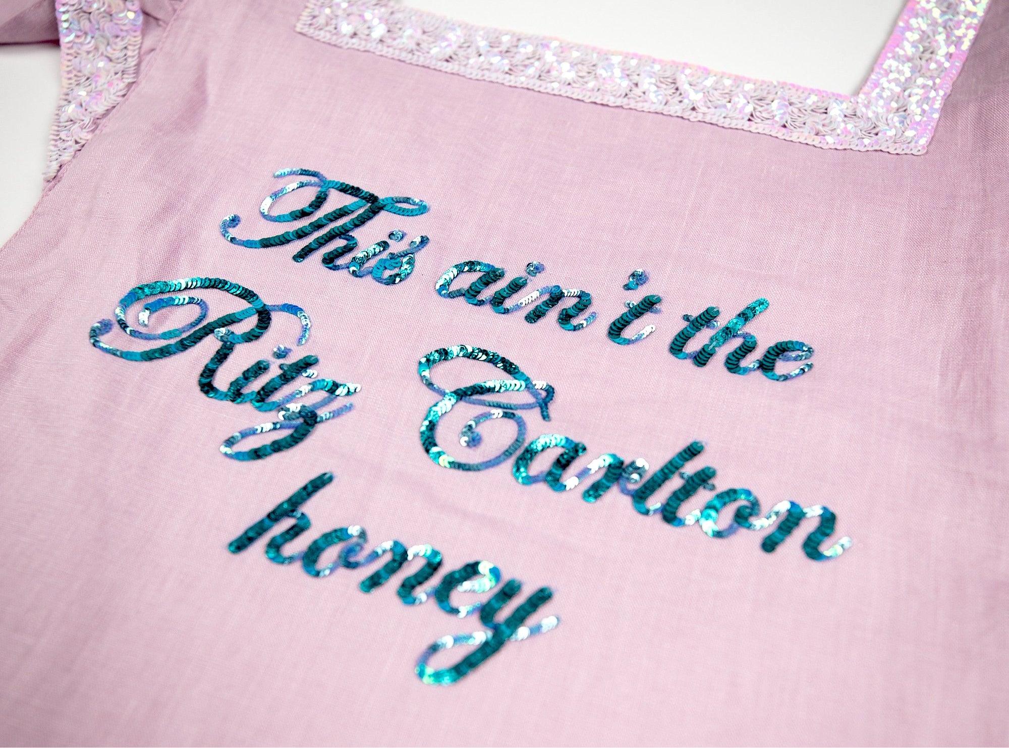 I made a fucking apron just for YOU!!! "This Ain't The Ritz Carlton Honey." This kitchen ain't a damn hotel. Kick your feet up and pour yourself a DRINK!! This apron is GORGEOUS. It's sequined, ruffled and trimmed with iridescent white sequin lace!!! I love it!! Get you one before it's gone... this baby is super limited!! *One size fits all.. This apron is hand embroidered pink linen with hand sequined details. Dry Clean Only, Lightweight & Lined Linen Fabric, Ashley Longshore Signature Loop Tag.